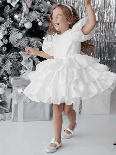 Smiling girl in white ruffled dress dancing by Christmas tree with silver gifts, FashionRay