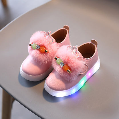 Pink kids' fashion sneakers with fluffy pom-poms, jeweled crown décor, and LED light soles on chair