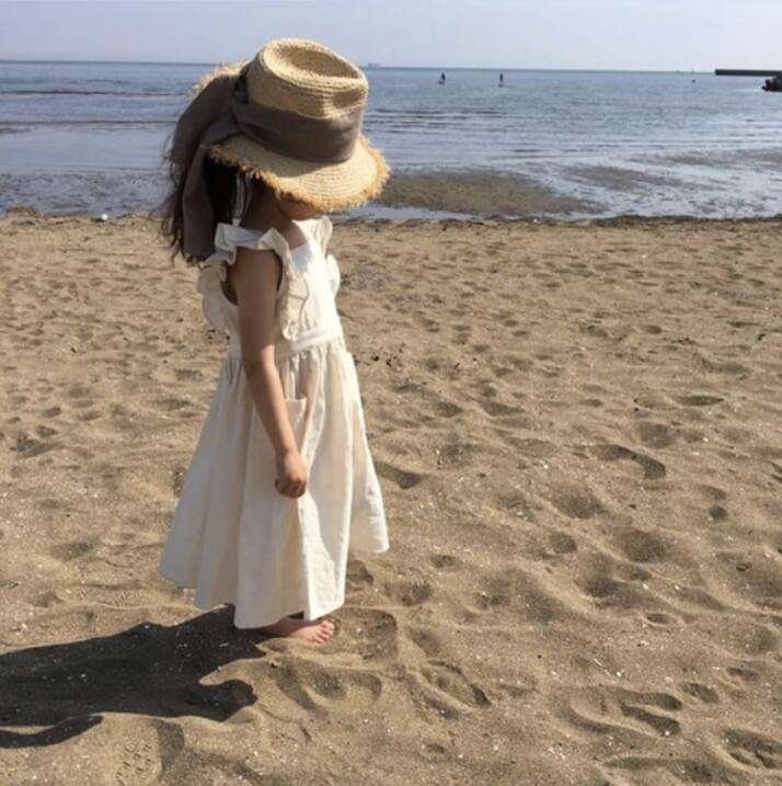Young girl in a cream dress and straw hat standing barefoot on a sandy beach by the sea