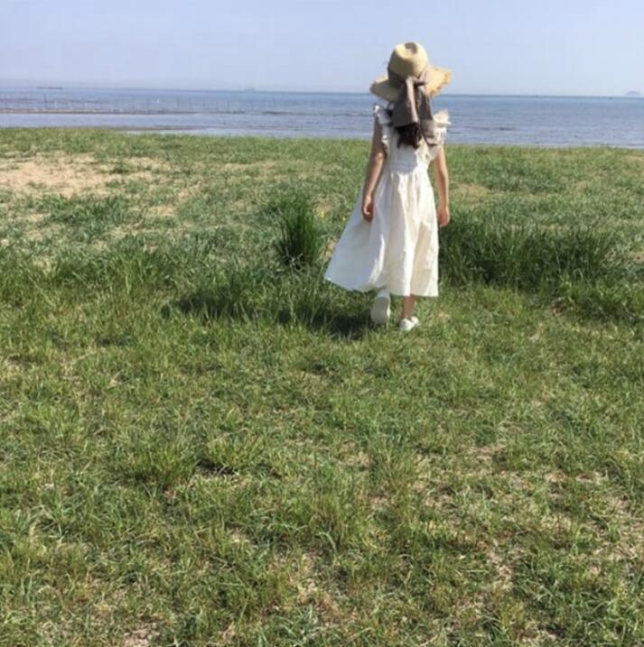 Woman in white dress and straw hat standing on grassy field by the sea, FashionRay style