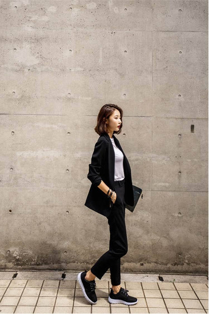 Woman in stylish black blazer, white top, and sneakers walking by concrete wall, FashionRay