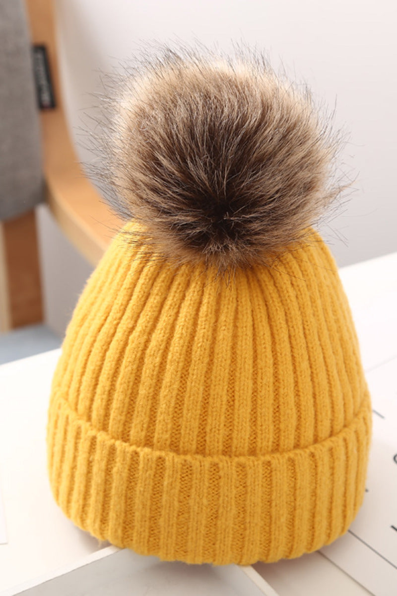 Yellow knit beanie with faux fur pom-pom on table, trendy winter hat from FashionRay