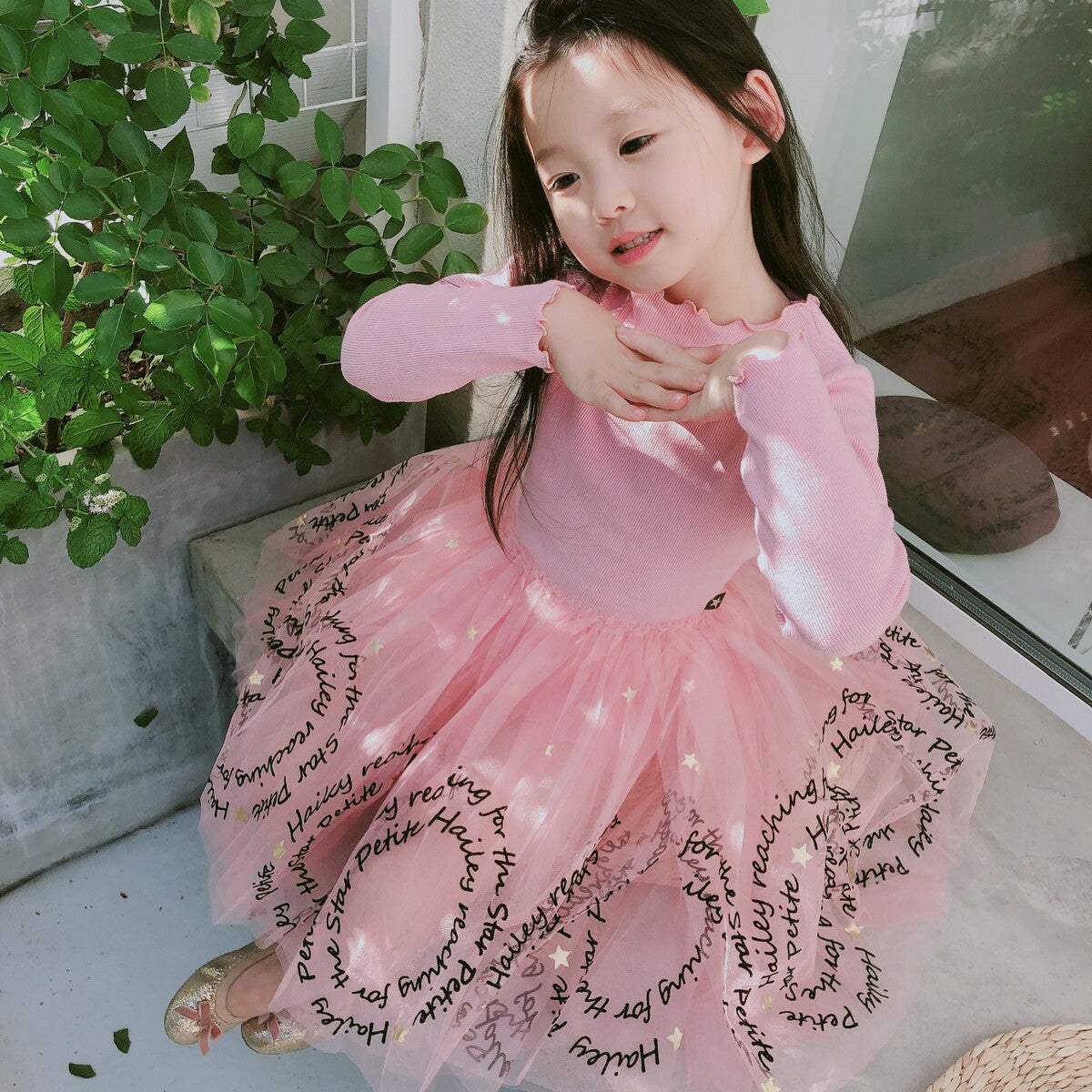 Young girl in a pink tulle dress with script text, posing near greenery, FashionRay style