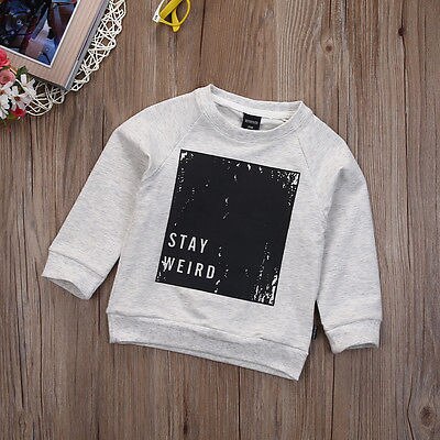 Grey kids sweatshirt with 'STAY WEIRD' graphic on wooden background, FashionRay style