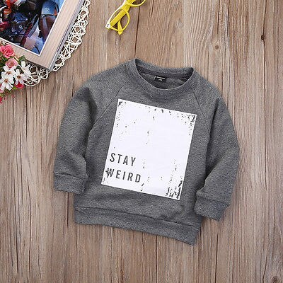 Gray toddler sweatshirt with 'STAY WEIRD' print, laid on wood, showcased by FashionRay.