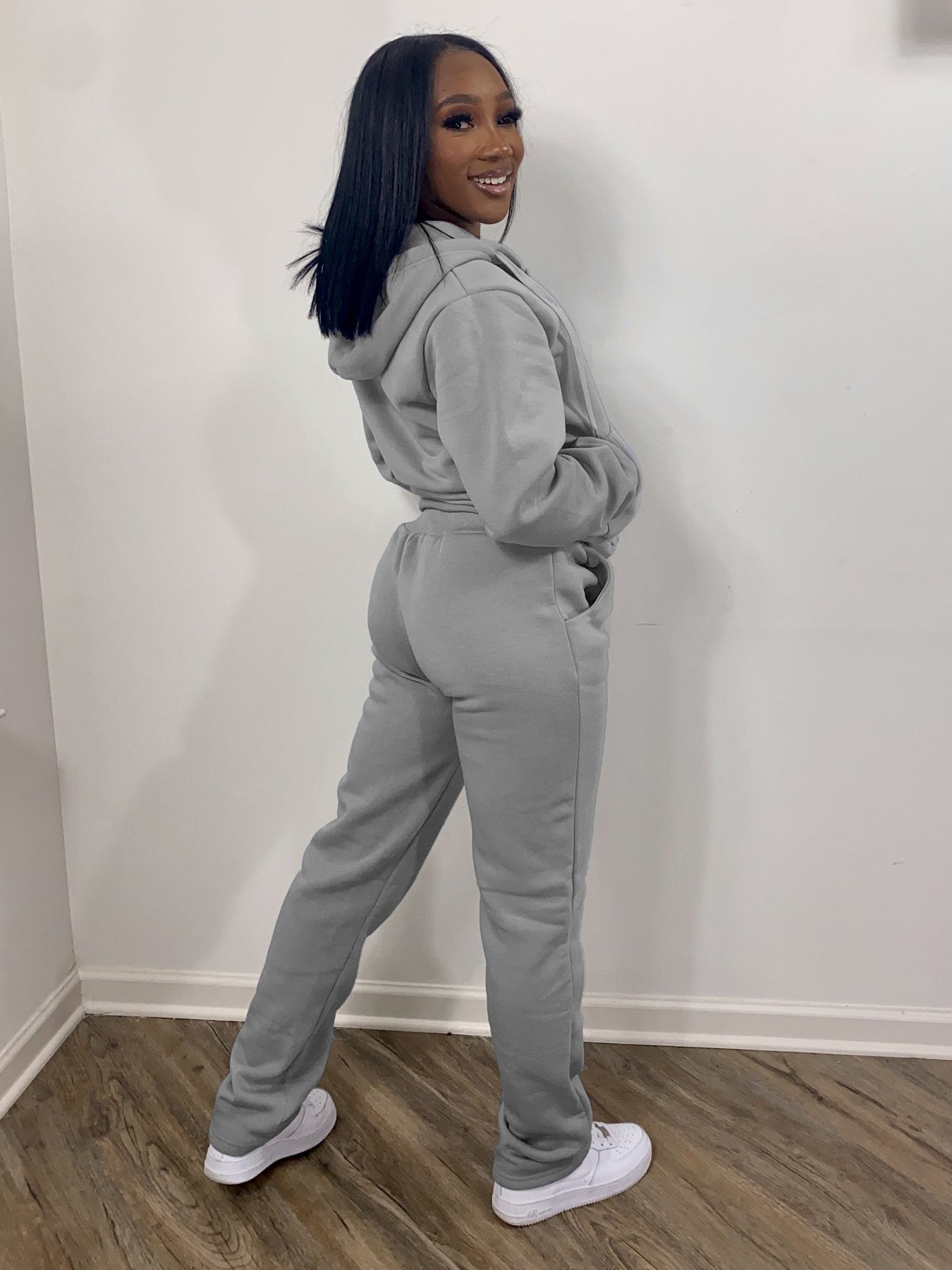 Woman modeling a light gray hoodie and sweatpants set with white sneakers on wood floor, FashionRay style