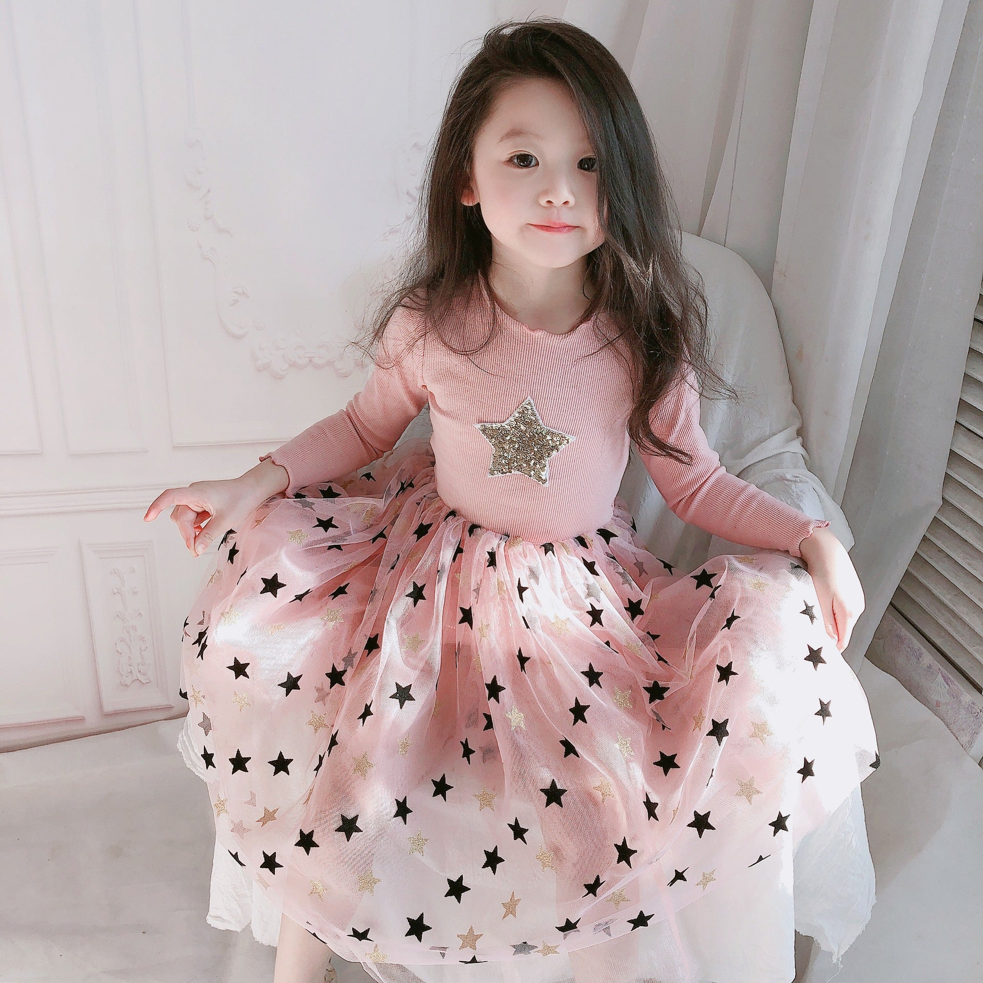 Girl in pink star-patterned dress sitting indoors, trendy kids fashion at FashionRay