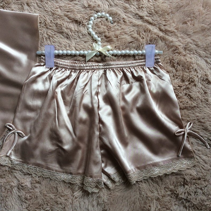 Silky beige satin shorts with lace trim and bow detail, displayed on a plush surface at FashionRay.