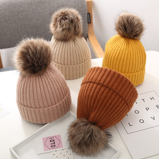Knit beanies with faux fur pom poms in beige, pink, mustard, and brown on a white table, FashionRay