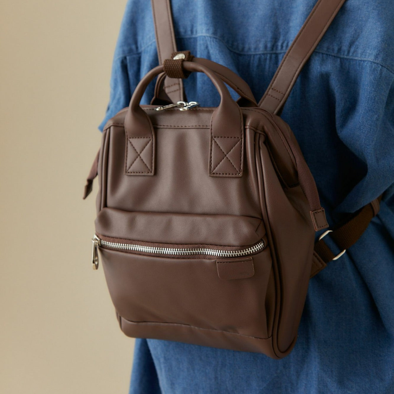 Brown leather mini backpack with front zipper pocket, worn over blue denim shirt, FashionRay style