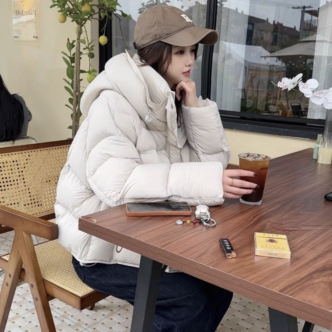 Woman in beige puffer jacket and cap at café table with iced coffee, FashionRay style