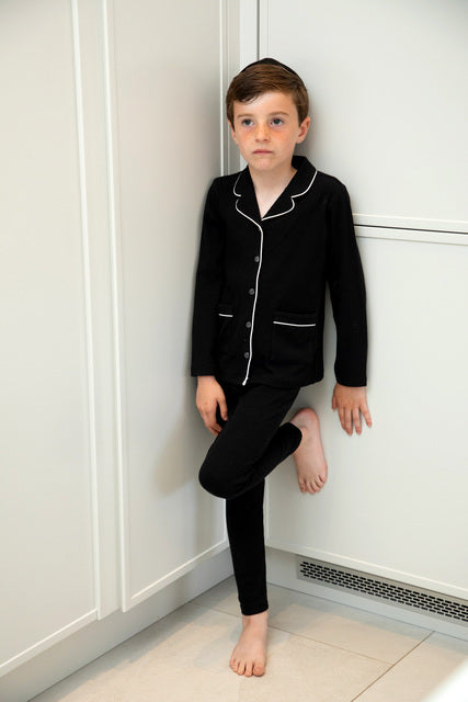 Boy in black pajama set with white piping, standing indoors by white cabinets, FashionRay style.