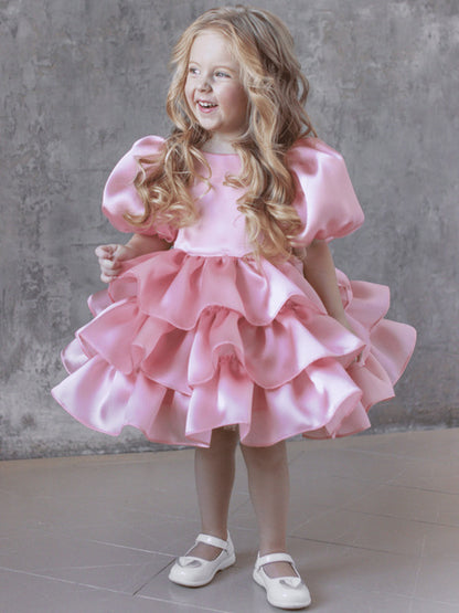 Smiling young girl in a ruffled pink dress and white shoes, posing indoors, FashionRay style
