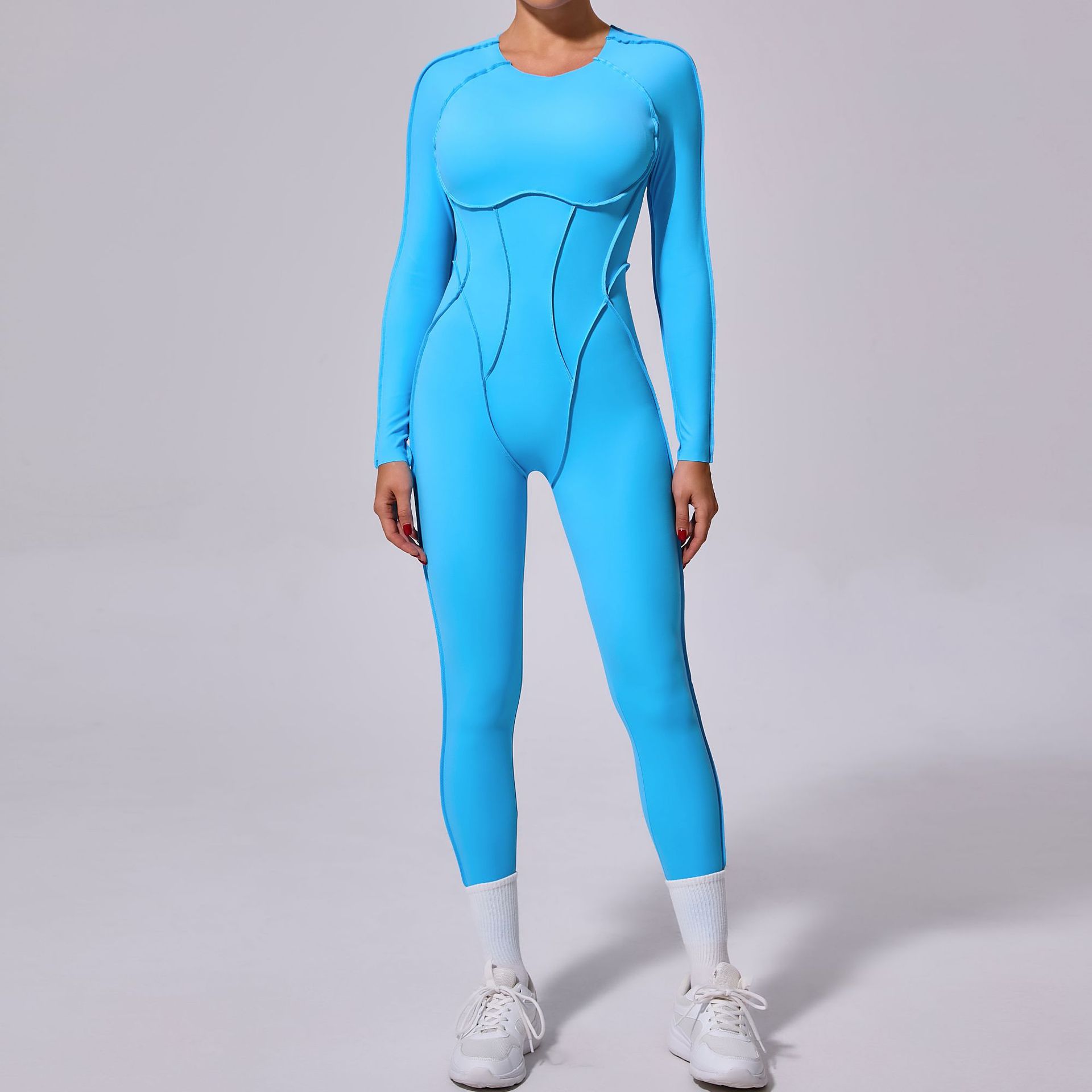 Woman modeling a bright blue long sleeve jumpsuit with sporty white sneakers on a plain background.