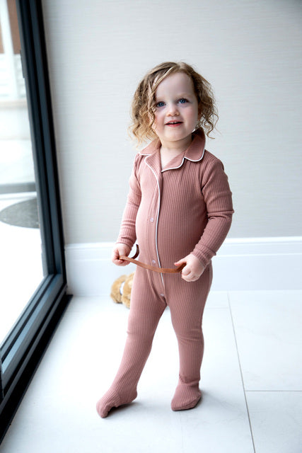 Smiling toddler in a pink ribbed jumpsuit by window, showcasing FashionRay comfort fashion