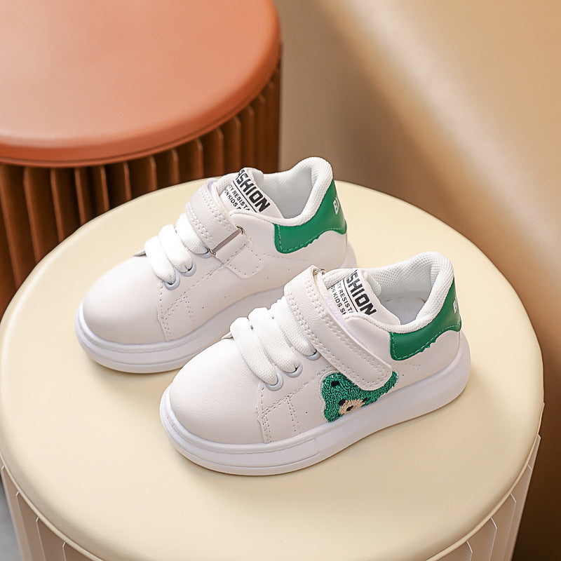White kids' sneakers with green accents and velcro straps on a beige ottoman, FashionRay style