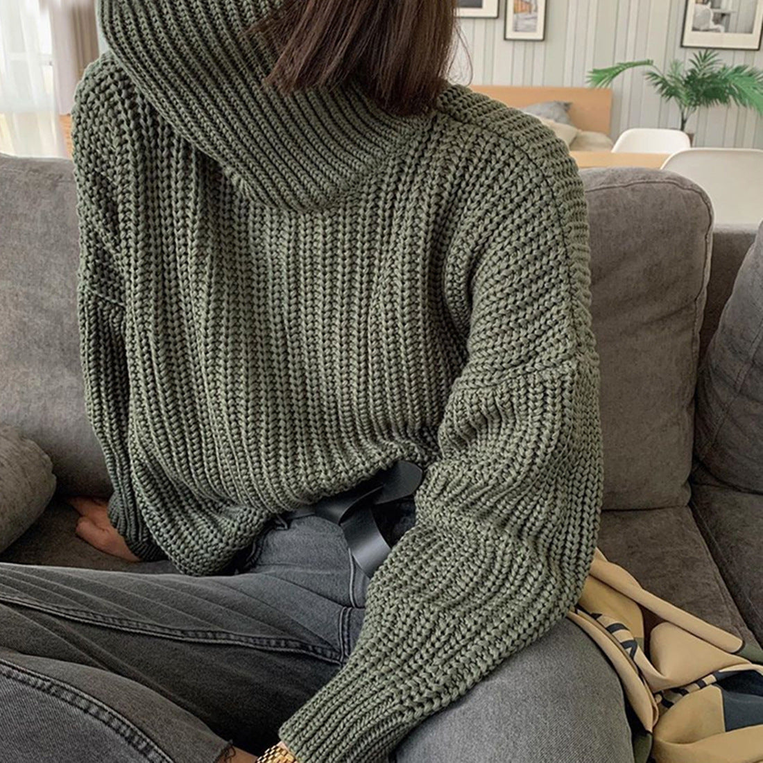 Woman in chunky olive green turtleneck sweater and gray jeans sitting on sofa, FashionRay style