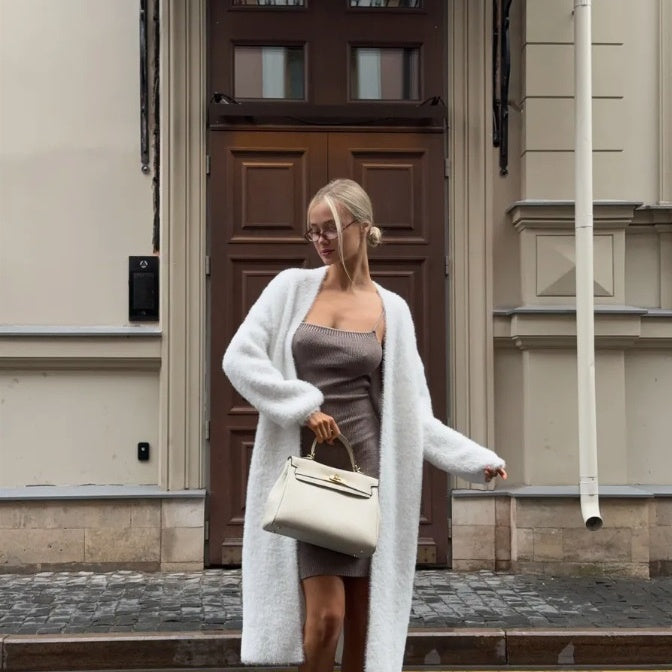 Trendy woman in white cardigan and taupe dress holding white handbag outside urban building