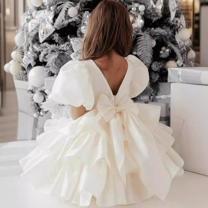Girl in ruffled white dress with bow sitting by decorated Christmas tree, FashionRay style