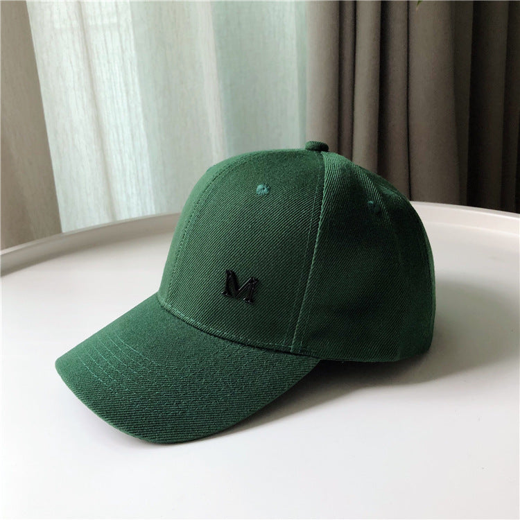 Green baseball cap with black M logo on brim, placed on white table, FashionRay style