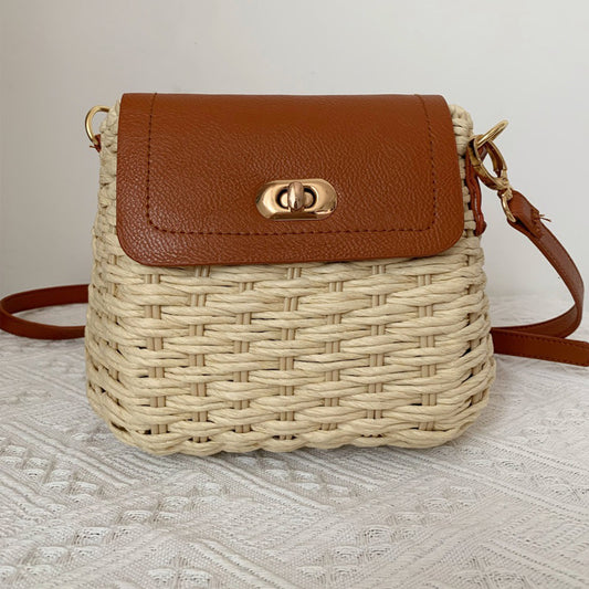 Woven straw crossbody bag with brown leather flap and gold clasp on white textured surface