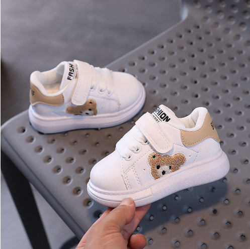 White toddler sneakers with bear patch and beige accents on perforated gray chair, FashionRay