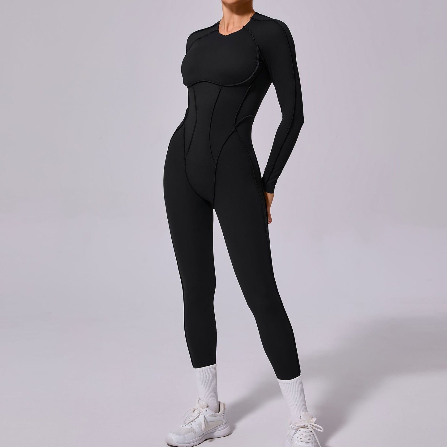 Woman wearing black long sleeve fitted jumpsuit with white sneakers on plain background