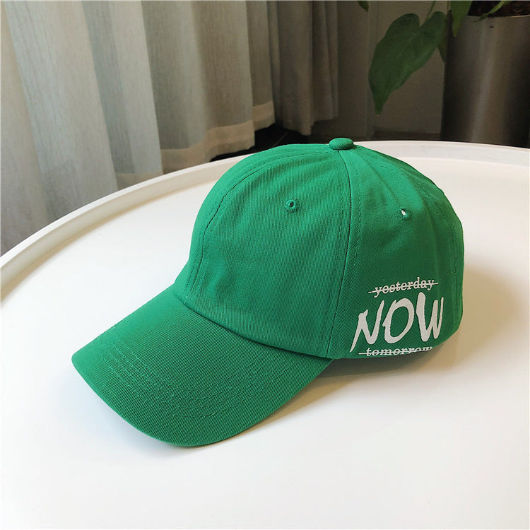 Green FashionRay baseball cap with bold NOW text, displayed on a white table indoors